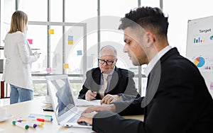 Focus on Business manager wearing eyeglass writing on paper and staff sitting typing and seeing on laptop near colour pens. Woman