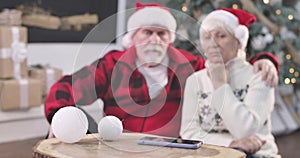 Focus changes from smartphone to faces of sad senior couple sitting next to Christmas tree. Retirees waiting for call