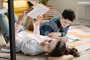 Focus of cheerful child lying on floor with book near brother writing in notebook