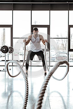 Focus of concentrated sportsman exercising with battle ropes in modern gym