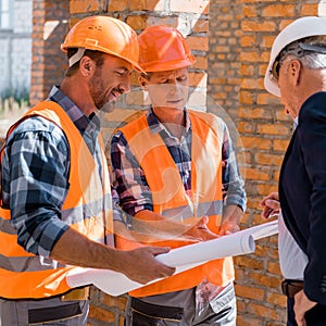 Focus of constructors near mature businessman in helmet looking at blueprint