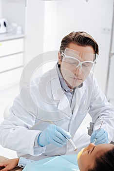 Focus of dentist in face shield and latex gloves holding dental instruments near african american woman