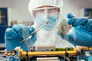 Focus of engineer holding small stone with tweezers and microchip near computer motherboard