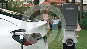 Focus EV station recharging battery for EV car on blurred family. Fastidious