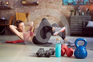 Focus on fitness equipments, barbell and kettlebell. Woman doing sit-ups in the background. Concepts about home workout, fitness,