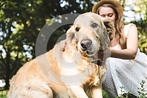 Focus of girl in white dress and straw hat petting golden retriever while sitting on meadow with closed eyes