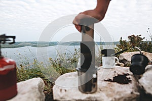 Focus on grass on cliff and blurred image of traveler pressing aeropress on metal mug on cliff at lake, brewing alternative coffee