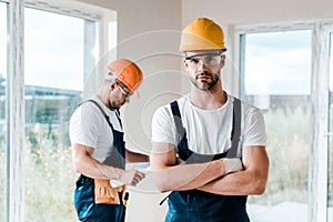 Focus of handsome handyman in goggles standing with crossed arms near coworker
