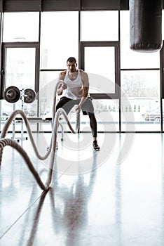 Focus of handsome sportsman exercising with battle ropes in modern gym