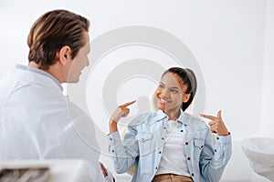 Focus of happy african american woman pointing with fingers at braces near orthodontist in clinic