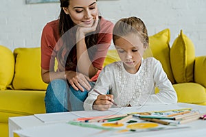 Focus of happy babysitter sitting on yellow sofa near kid drawing in living room