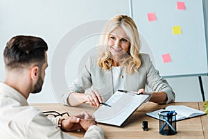 Focus of happy recruiter holding pen near clipboard and looking at bearded man
