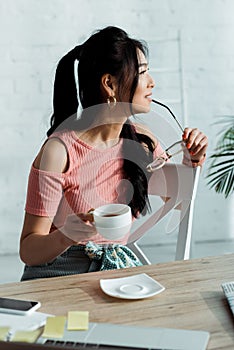 Focus of happy young asian woman holding gasses near lips and cup of tea