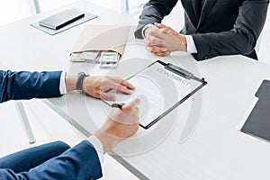Focus of man holding clipboard and pointing with finger at contract with pen near business partner gesturing in office