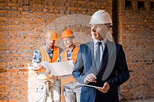 Focus of mature businessman holding digital tablet near constructors