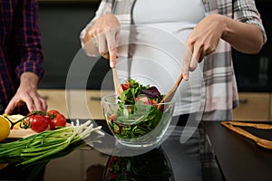 Focus on pregnant woman`s hands holding wooden spoons and mixing ingredients in a glass bowl, preparing delicious healthy salad