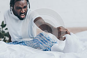 Focus of screaming african american man suffering from foot pain in bedroom