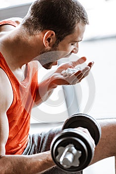 Focus of sportsman with opened mouth exercising with dumbbell and looking at probiotic pills in gym