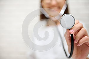 Focus on stethoscope phonendoscope in the hand of a blurred female doctor smiling toothy smile against white wall background with