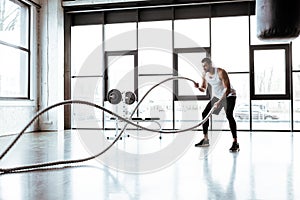 Focus of strong sportsman exercising with battle ropes in modern gym