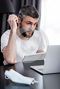 Focus of teleworker looking at laptop while holding notebook and pen near medical mask on table