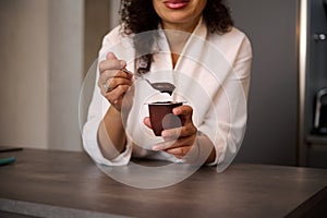 Focus on woman& x27;s hands, holding a teaspoon and a container with wholesome chocolate vegetarian yoghourt for