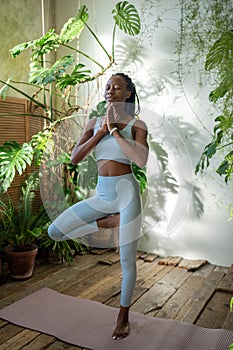 Black woman meditating in yoga tree pose stand on one leg with closed eyes at home garden. Wellness.
