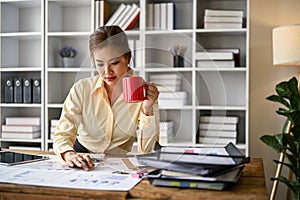 Focused Asian female accountant sipping coffee and analyzing business financial data on reports