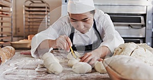 Focused asian female baker working in bakery kitchen, cutting dough for rolls in slow motion