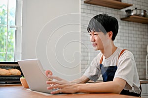 Focused Asian man in apron using laptop in his kitchen, joining online cooking class