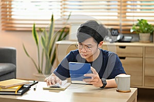 Focused asian man calculating household finances or taxes on calculator, sitting in bright living room