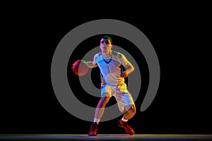 Focused athletic man, basketball player dribbles ball with one hand against black studio background in mixed neon light.