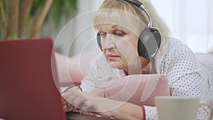 Focused beautiful senior woman messaging online typing on laptop keyboard. Caucasian concentrated retiree in headphones