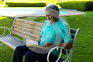 Focused biracial senior man wearing blue t-shirt reading book while sitting on bench in park