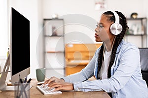 Focused black lady student with headphones works on computer for online class