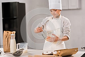 focused boy in chef hat and apron whisking eggs in bowl at table