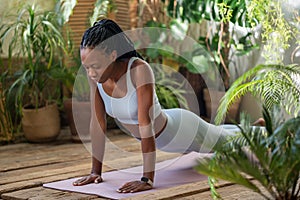 Focused on breath active black woman doing plank on fitness mat with closed eyes in room with plants