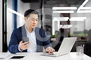 Focused businessman in office using laptop and smartphone with serious expression