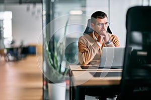 Focused businessman sitting at desk with laptop, thoughtfully analyzing data or planning strategy in a modern office