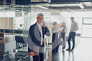 Focused businessman is talking phone with client while standing in office on colleagues background