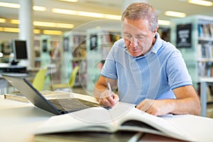 Focused casual mature male writing in notepad and reading books in the library