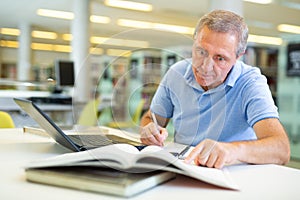 Focused casual mature male writing in notepad and reading books in the library