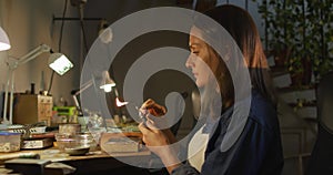 Focused caucasian female jeweller sitting at desk, making jewelry in workshop