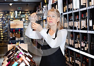 Focused mature woman wine producer inspecting quality of wine in wineshop on background with shelves of wine bottles