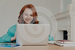 Focused cheerful redhead successful businesswoman does research, makes business project, sits in front of laptop computer at home