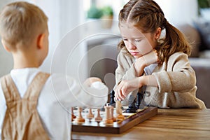 Focused children playing chess game at home while sitting in living room at table with chessboard