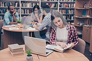 Focused concentrated attractive clever female student is learning with thesaurus in hard bookcase, wearing casual checkered shirt