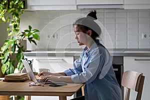 Focused concentrated young Asian female freelance blogger working on laptop in kitchen