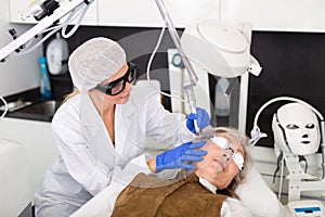 Focused cosmetologist performing laser resurfacing to senior woman