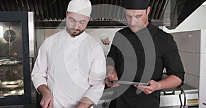 Focused cucasian male chef instructing trainee male chef using tablet in kitchen, slow motion